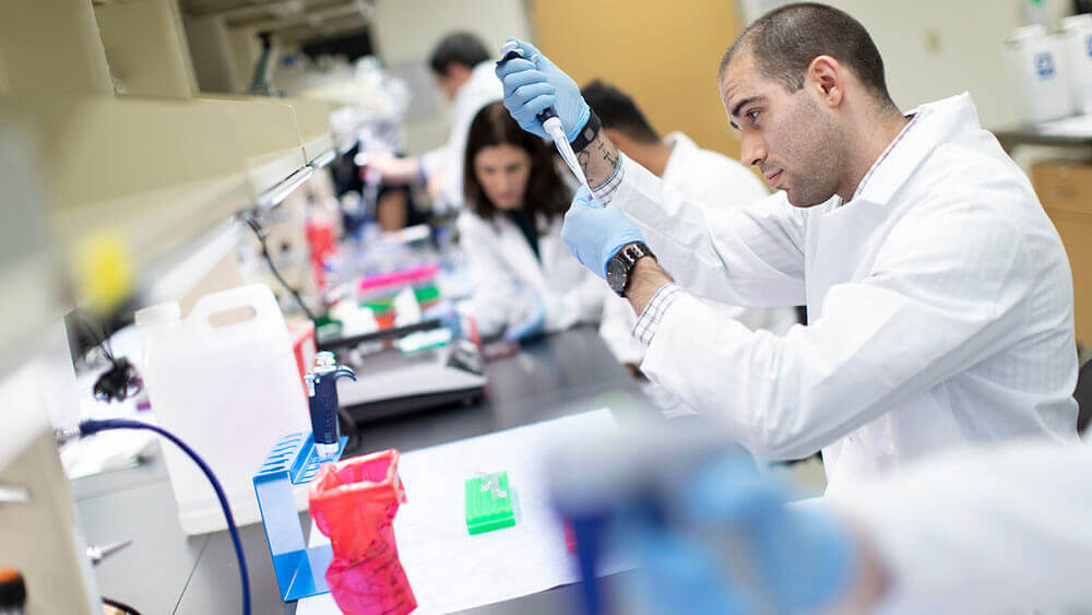 A medical student wearing a lab coat and gloves uses a pipette to process fluid in the research laboratory