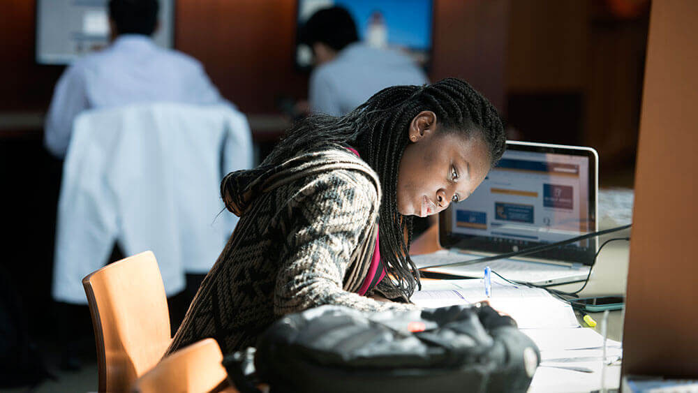 A student writes diligently in her notebook with her laptop open on a desk in the Netter Library