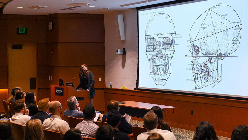 Medical student Katelyn Norman stands in front of a large display of a skull sketch as she presents her capstone findings